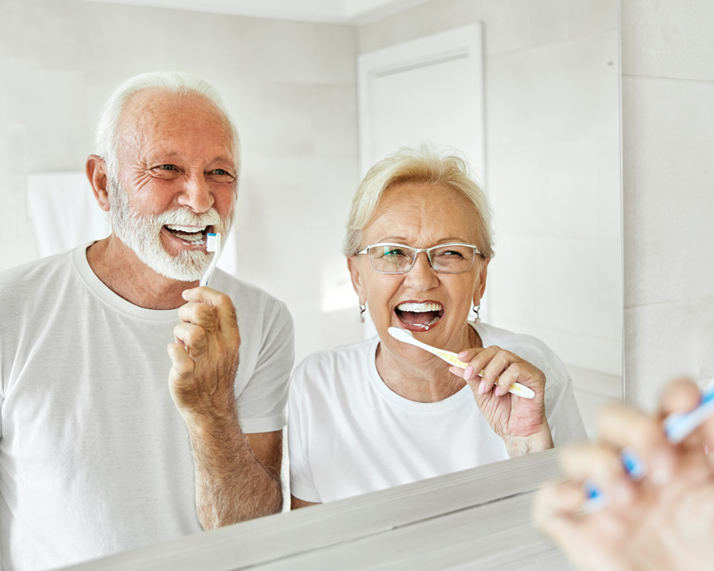 Older couple happily brushing their teeth showing the importance of routine cleanings